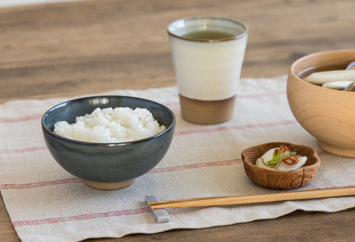 Bowl of rice, cup of tea, and small dish on a wooden table with a cloth.