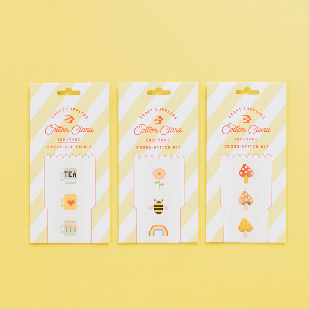 Three packages of Cotton Clara cross-stitch kits on a yellow background