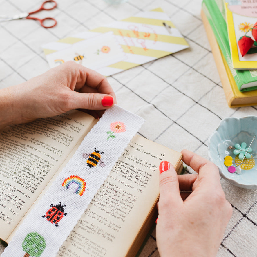 Person holding a cross-stitched bookmark with colorful designs on an open book.
