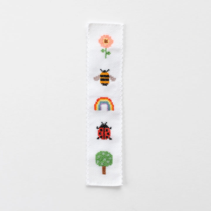 Cross-stitched bookmark with a flower, bee, rainbow, ladybug, and tree on a white background