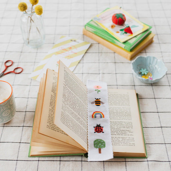 Open book with a cross-stitched bookmark featuring various designs on a checkered tablecloth.