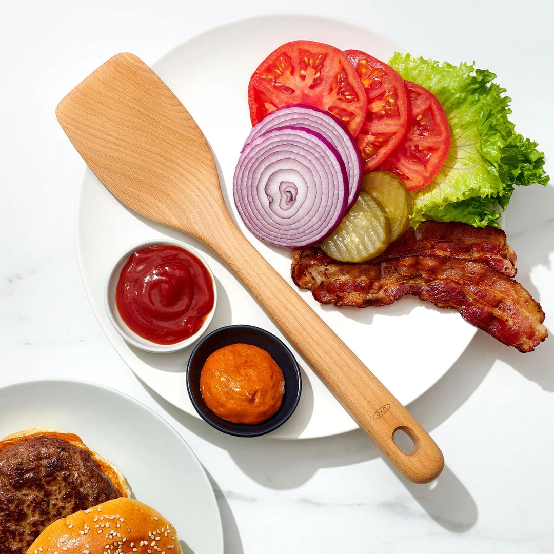 Hamburger ingredients including a wooden spatula, ketchup, mustard, tomatoes, onions, pickles, and bacon on a white background.