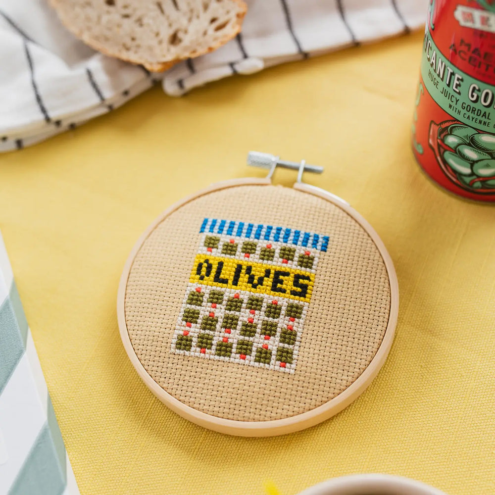 Cross-stitched hoop with 'OLIVES' text on a yellow surface with bread and a can.