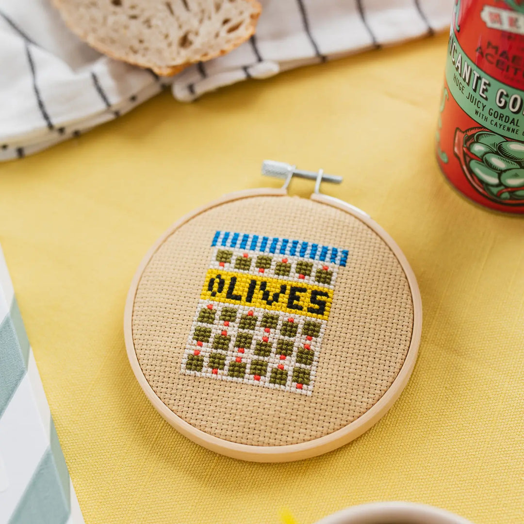 Cross-stitched hoop with 'OLIVES' text on a yellow surface with bread and a can.