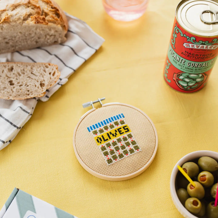 Cross-stitched hoop with 'OLIVES' design on a yellow table with bread and a can.
