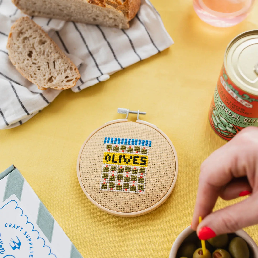 Cross-stitched hoop with 'OLIVES' text on a yellow table with bread and olive jar.