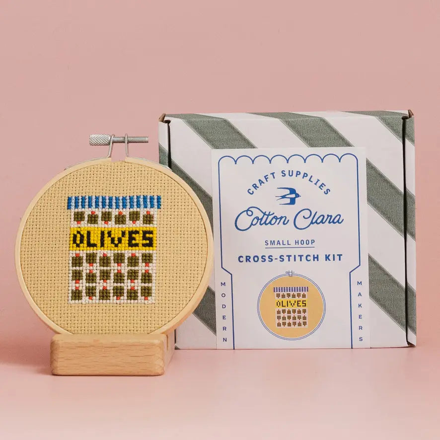 Cross-stitch kit and 'Olives' design next to Cotton Clara packaging on a pink background