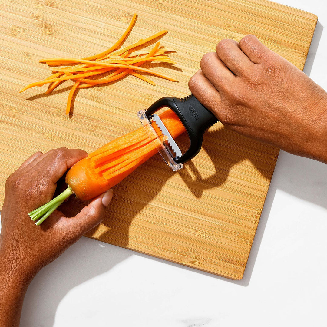Person using a carrot peeler on a carrot with sliced carrots on a wooden cutting board.