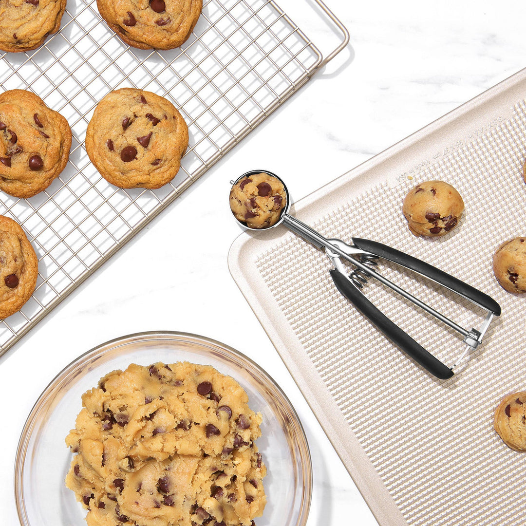 Chocolate chip cookies on a cooling rack, baking sheet, and plate with a cookie scoop.