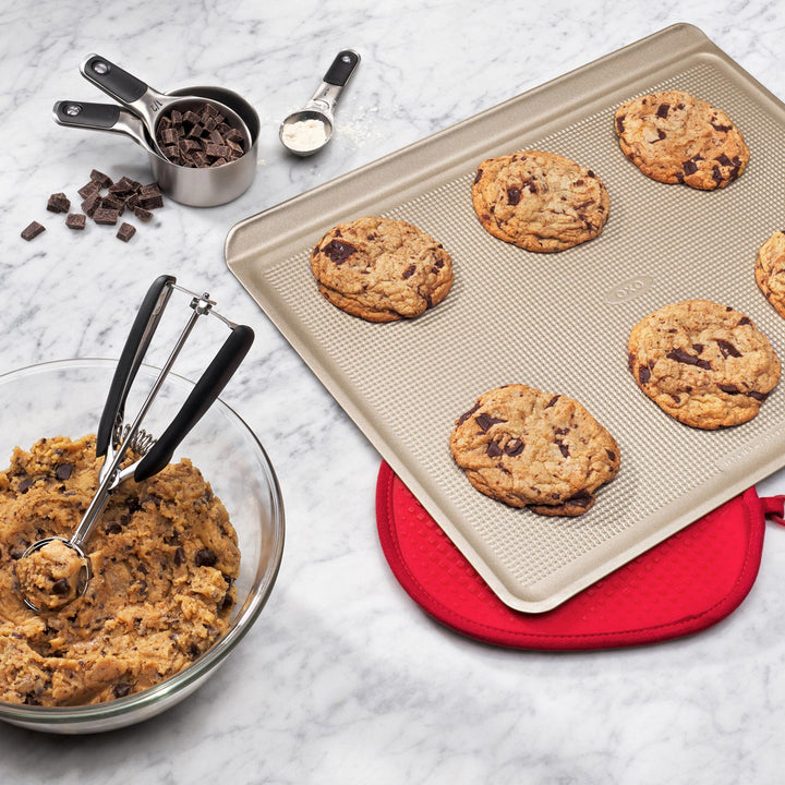 Baking tray with cookies, bowl of cookie dough, and measuring utensils on a marble surface