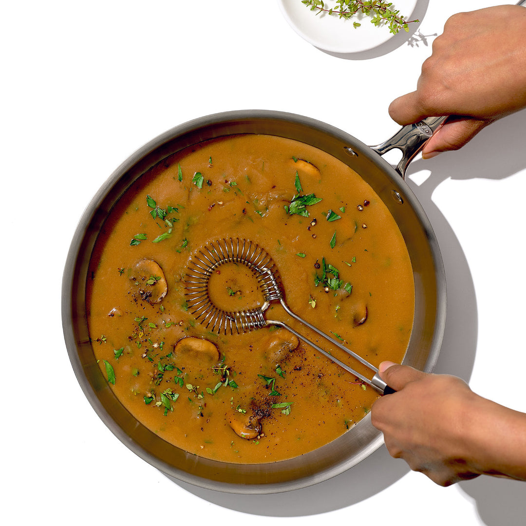 Person holding a sauce and gravy whisk over a pan of creamy mushroom soup with herbs.