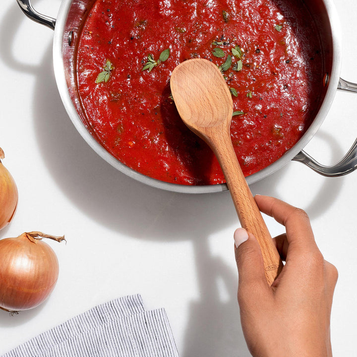 Red sauce in a pot with a wooden spoon, surrounded by onions on a white surface