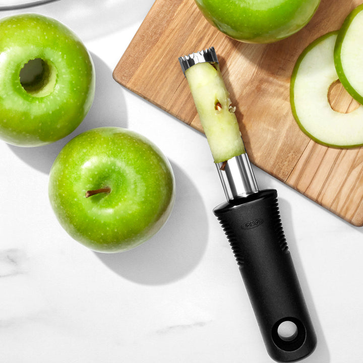 Green apples and an apple corer on a white surface with a wooden cutting board.