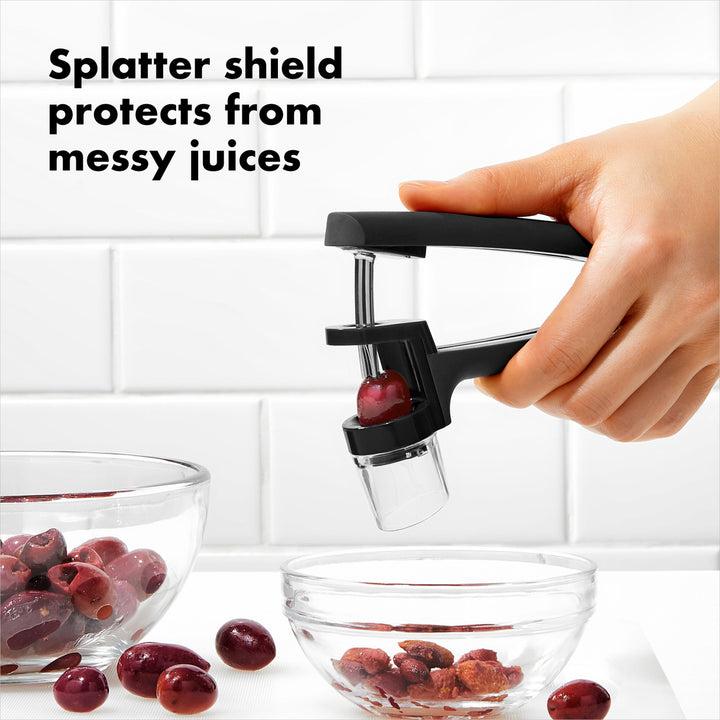 Hand using a cherry pitter with a splatter shield to remove pits from cherries, with text &