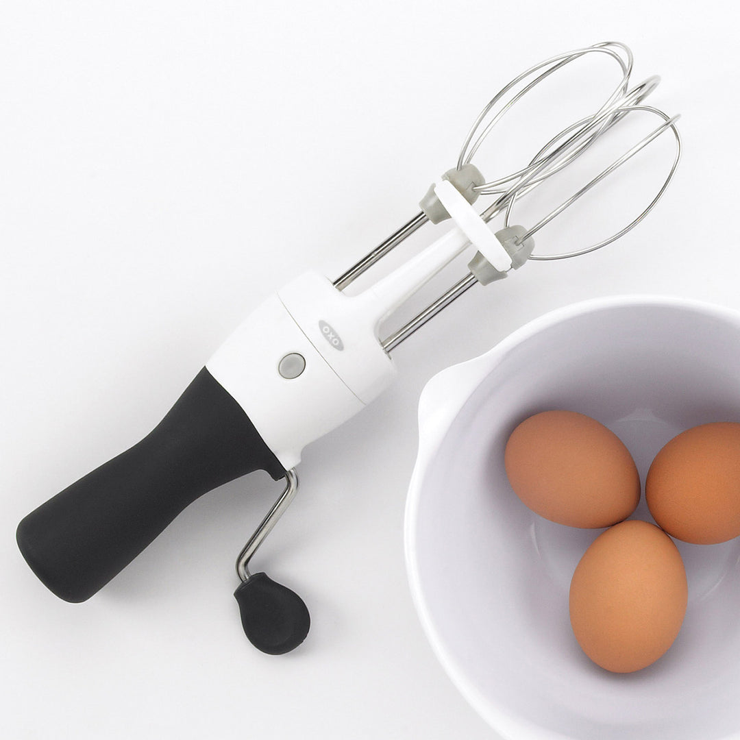 Handheld egg beater with whisk attachment next to a bowl of eggs on a white background