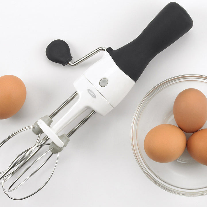 Handheld electric egg beater with black handle next to eggs on a white surface