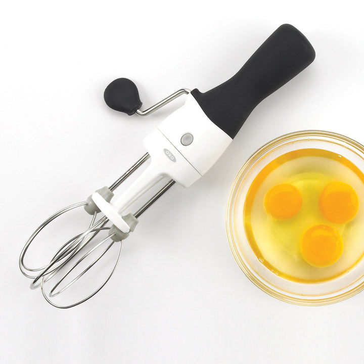 Handheld egg beater next to a bowl of eggs on a white background