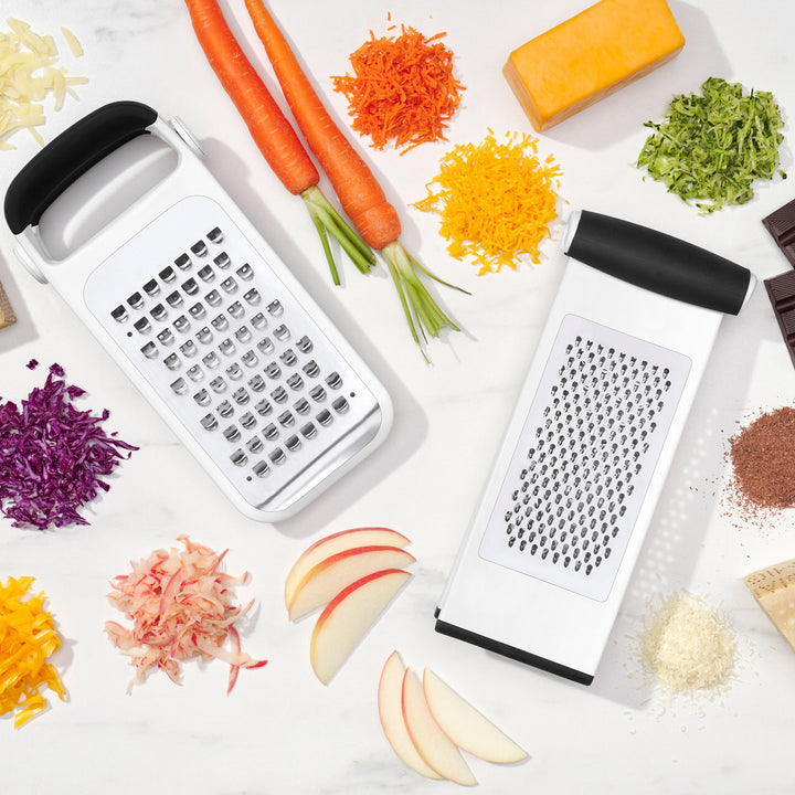 Two manual graters with grated vegetables and fruits on a white surface