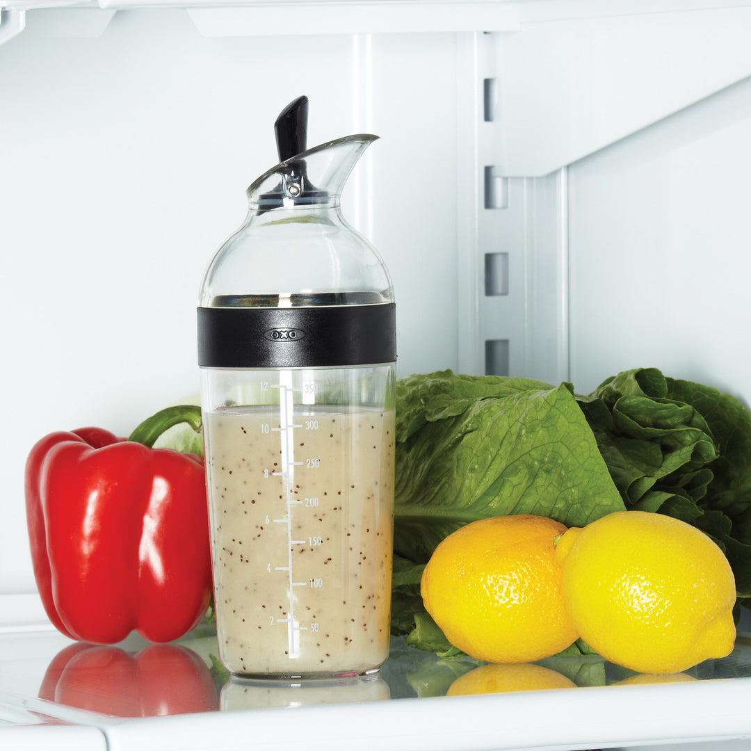 Clear bottle with black cap containing a liquid, surrounded by vegetables like red bell pepper, lemons, and lettuce inside an open refrigerator.