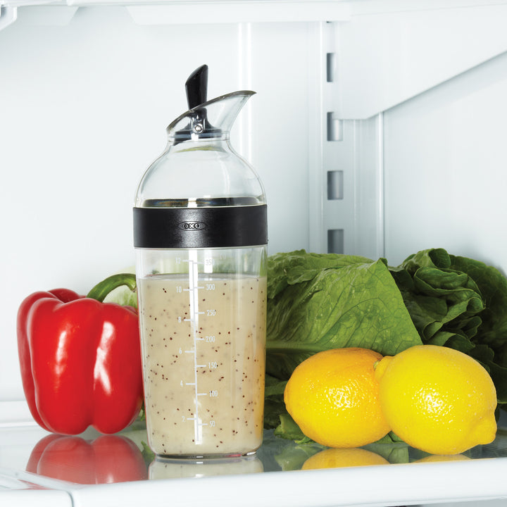 Clear bottle with black cap containing a liquid, surrounded by vegetables like red bell pepper, lemons, and lettuce inside an open refrigerator.