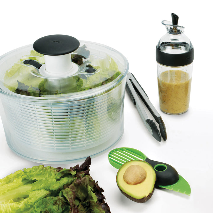 Salad spinner with salad, avocado, tongs, and dressing on a white background