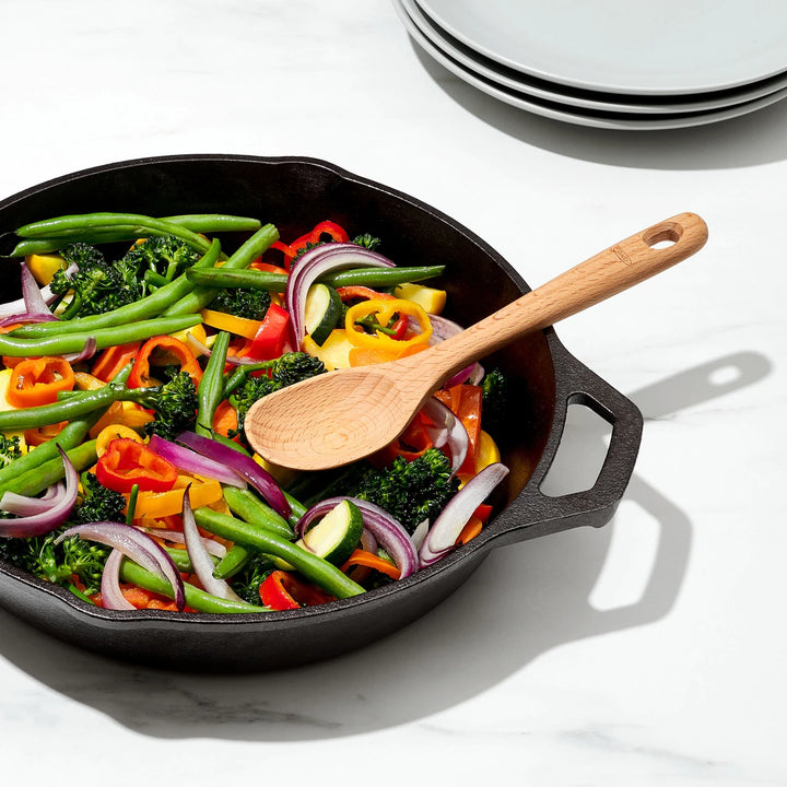 Black cast iron skillet with colorful vegetables and a wooden spoon on a white surface.