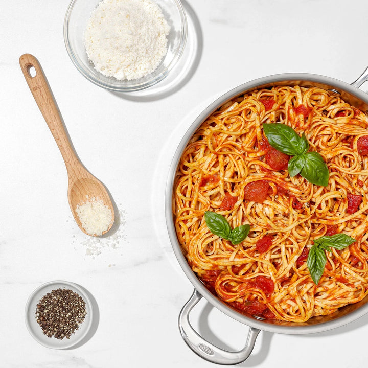 Pasta dish with tomatoes and basil in a pan on a white surface with a wooden spoon and small bowls of ingredients.