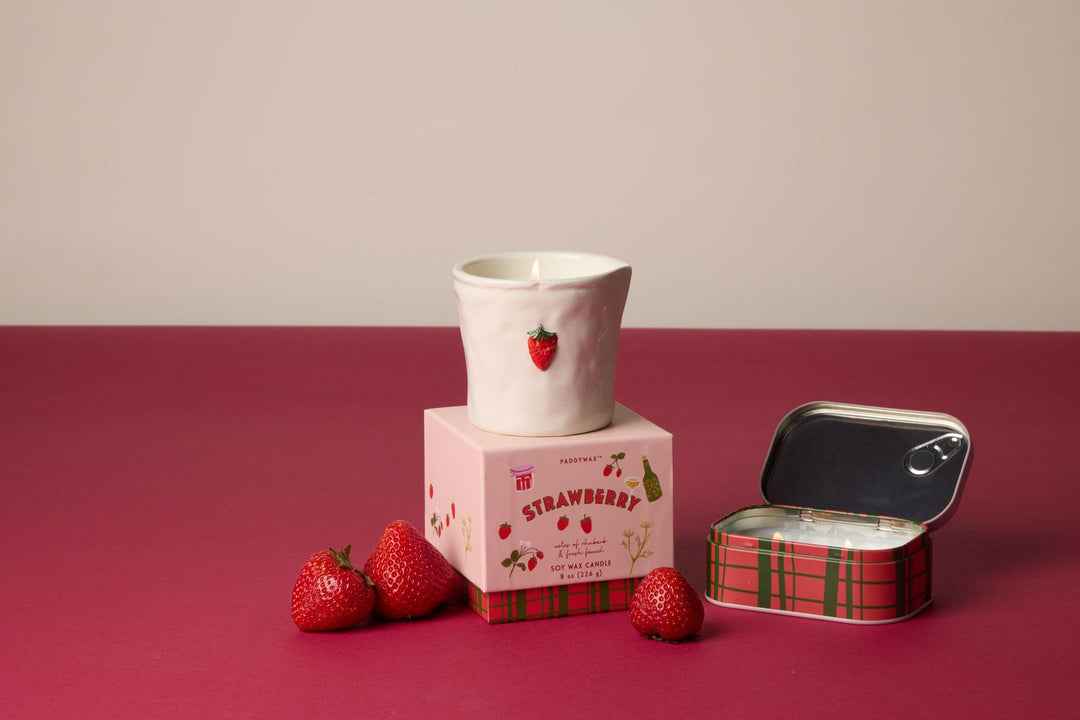 Candle with strawberry design, strawberry-themed packaging, and small tin on a red surface.