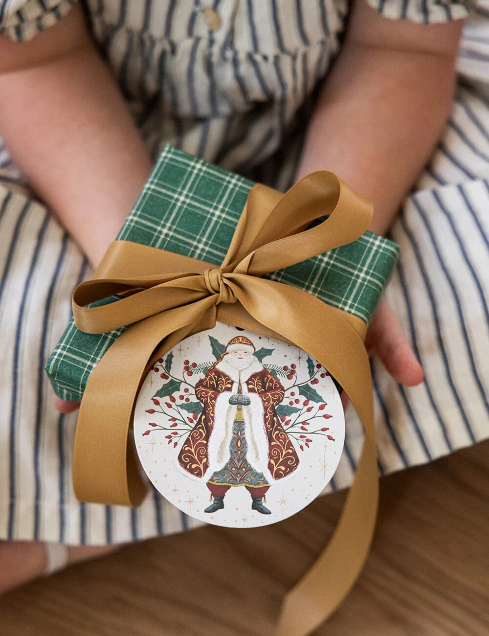 Gift wrapped in green paper with a brown ribbon held by a person wearing a striped shirt.