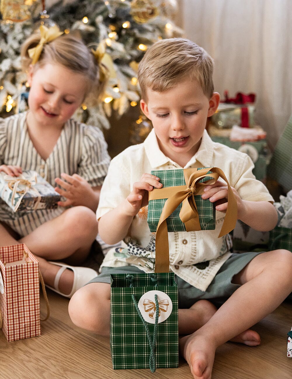 Two children sitting on the floor in front of a Christmas tree, opening presents.