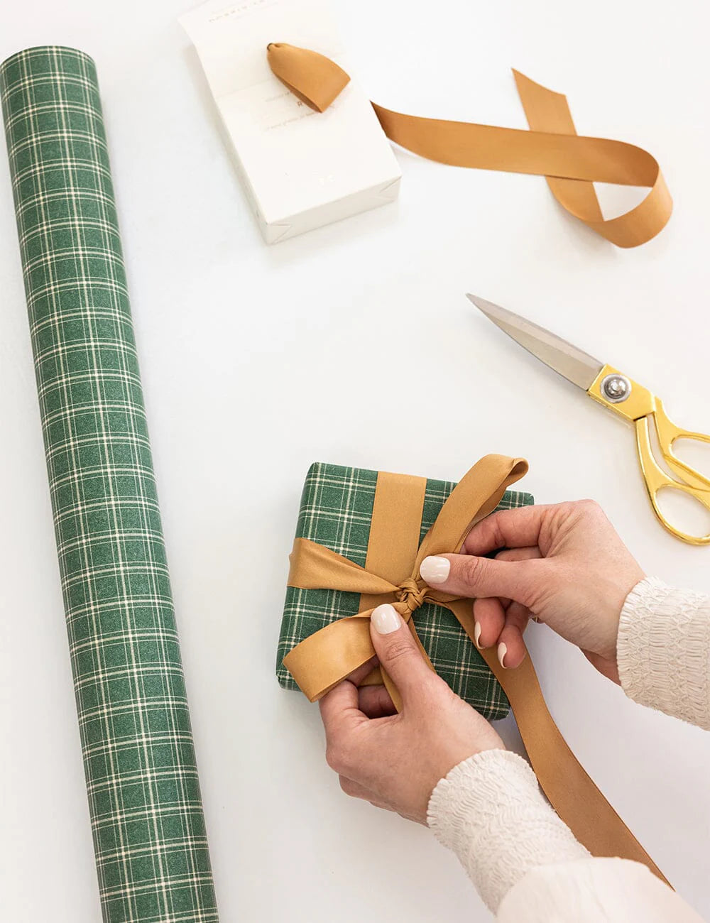 Person wrapping a gift with green plaid paper, brown ribbon, and scissors on a white surface.