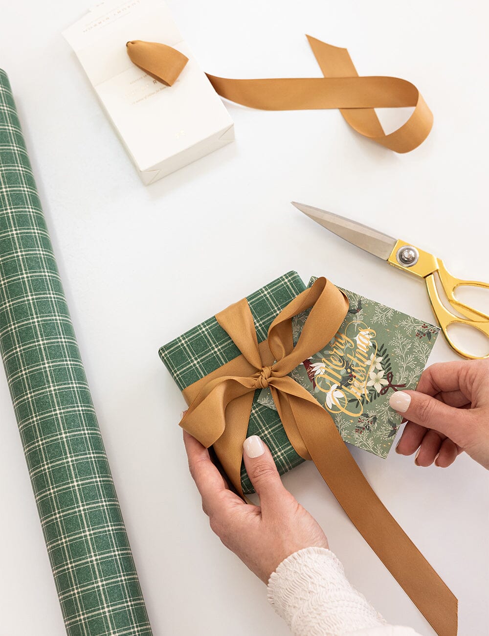 Person wrapping a gift with green paper and gold ribbon on a white surface.