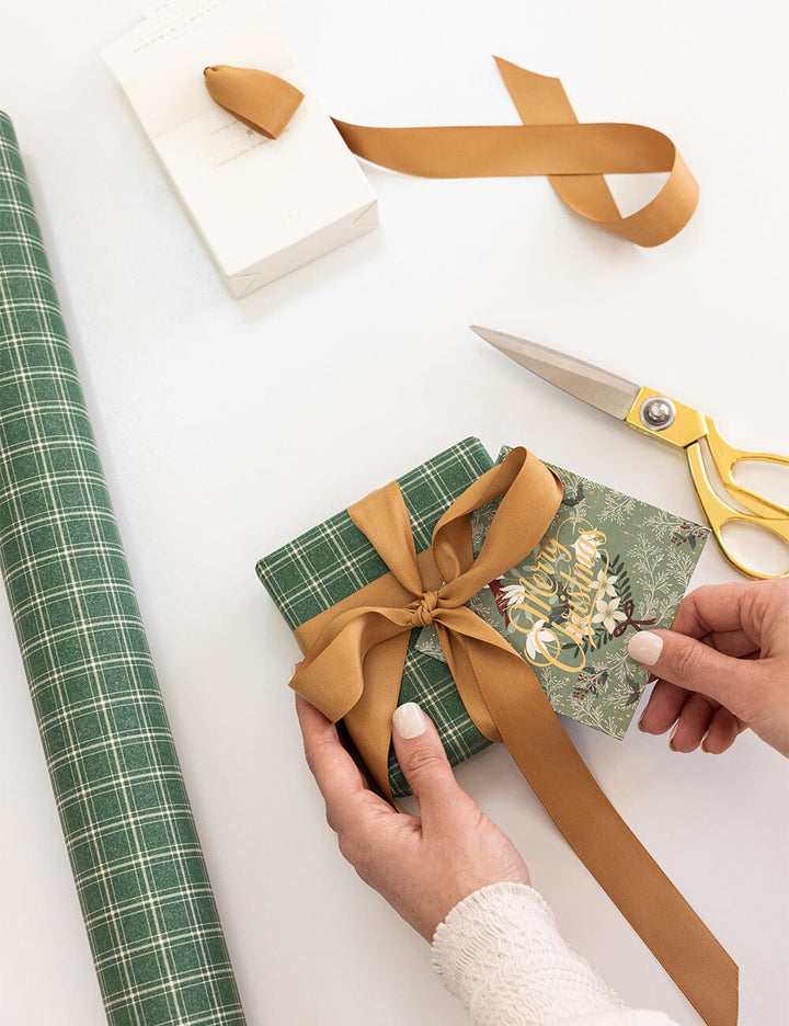 Person wrapping a gift with green paper and gold ribbon on a white surface.