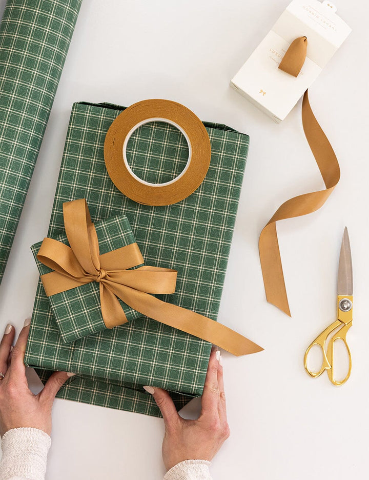 Green plaid gift box with a brown ribbon, tape, scissors, and additional wrapping paper on a white surface.