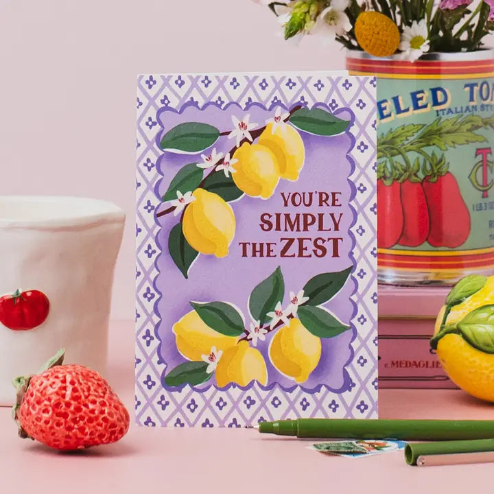 Notebook with lemon design and text 'You're Simply the Zest' on a pink surface with decorative items.
