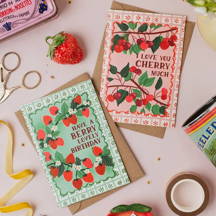 Two greeting cards with cherry designs on a table with stationery items.