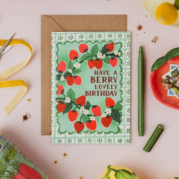 Greeting card with strawberry design and text 'Have a berry lovely birthday' on a pink surface with stationery items.
