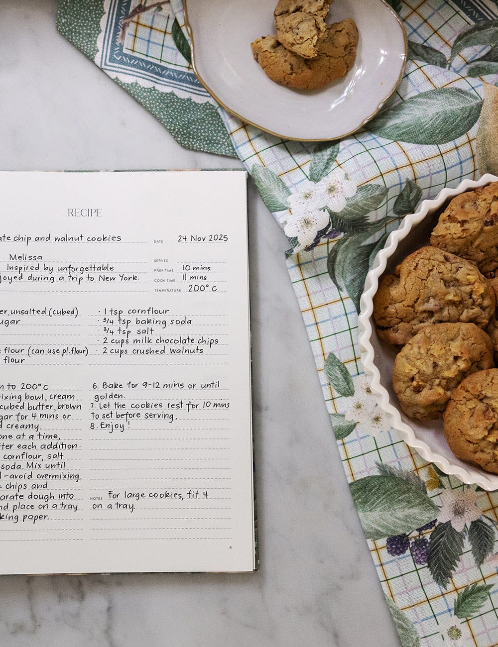 Baking ingredients and a recipe book on a marble surface with cookies.
