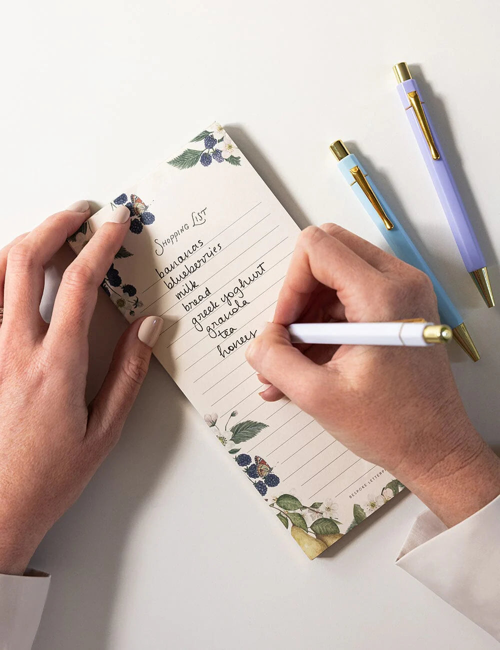 Person writing in a floral notepad with three pens on a white surface
