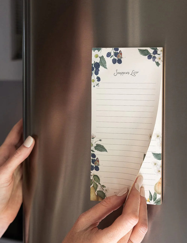 Person holding a floral-themed magnetic notepad against a refrigerator.