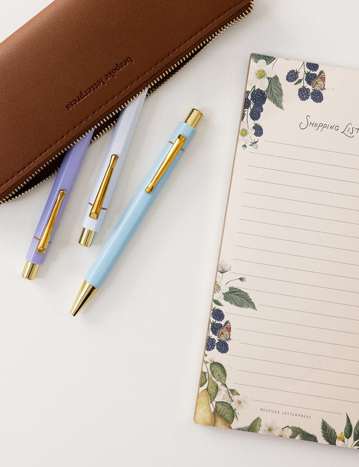 Three pens with gold accents on a white surface next to a floral notebook.