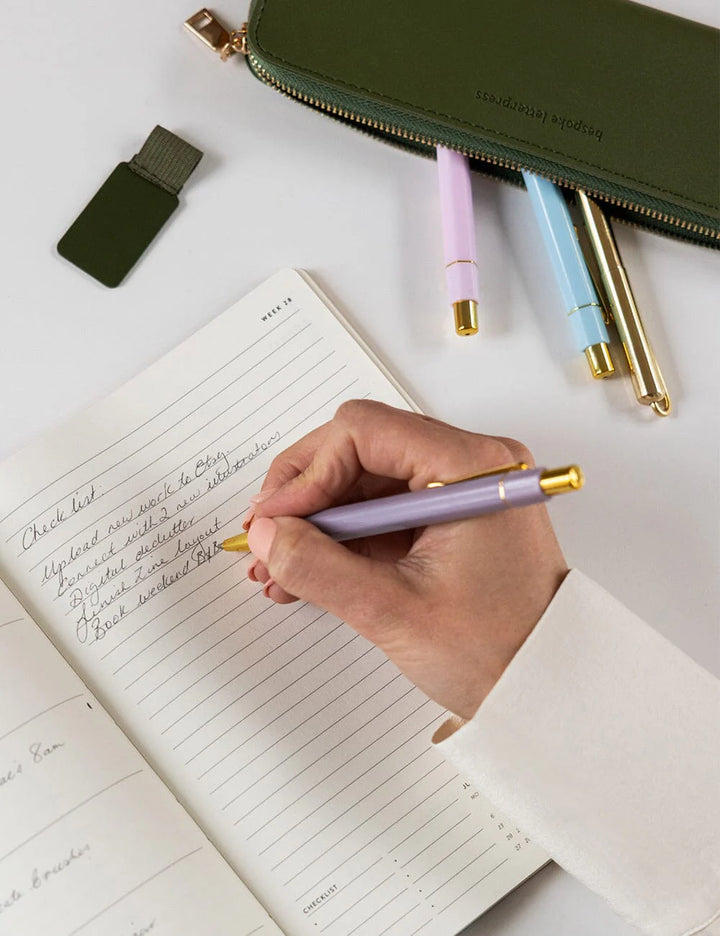Hand writing in a notebook with a pen, surrounded by more pens and a green case on a light surface.