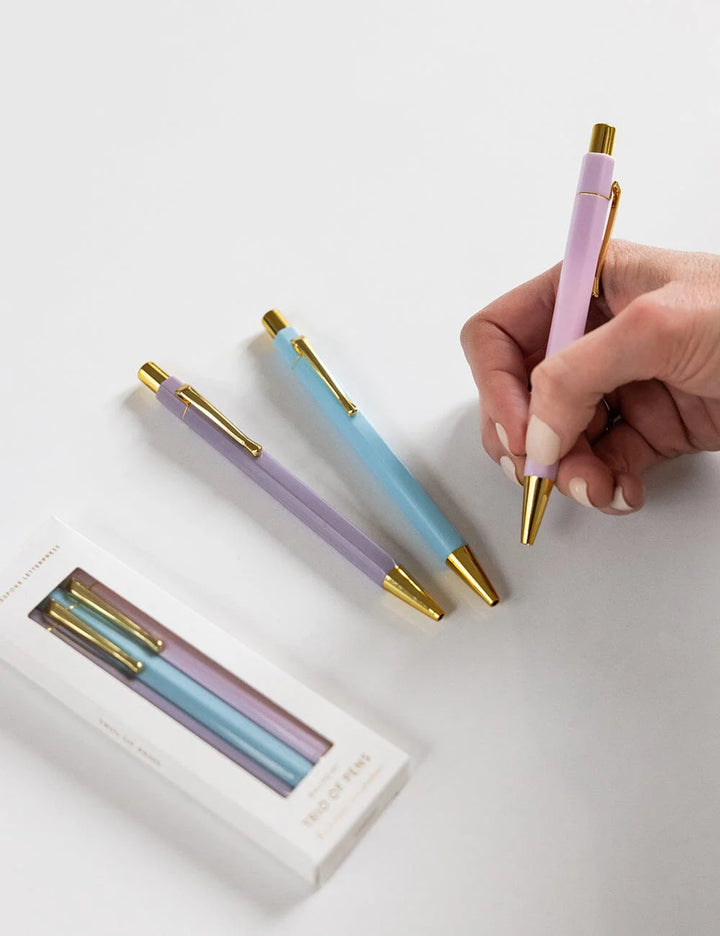 Set of pastel-colored pens with gold accents on a white surface, one pen held by a hand.