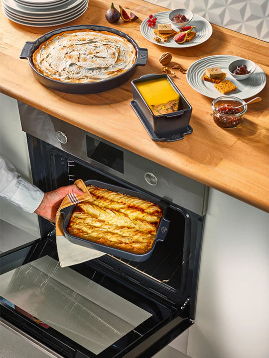 Person taking a dish out of an oven with a wooden table and food items in the background
