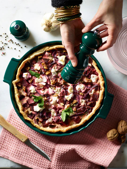 Person seasoning a quiche with a pepper grinder on a pink cloth