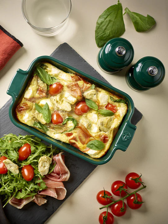 Green baking dish with a baked dish, tomatoes, and salad on a beige surface.