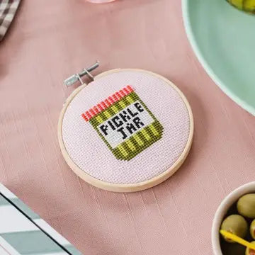 Cross-stitched pickled jar design on a pink fabric background