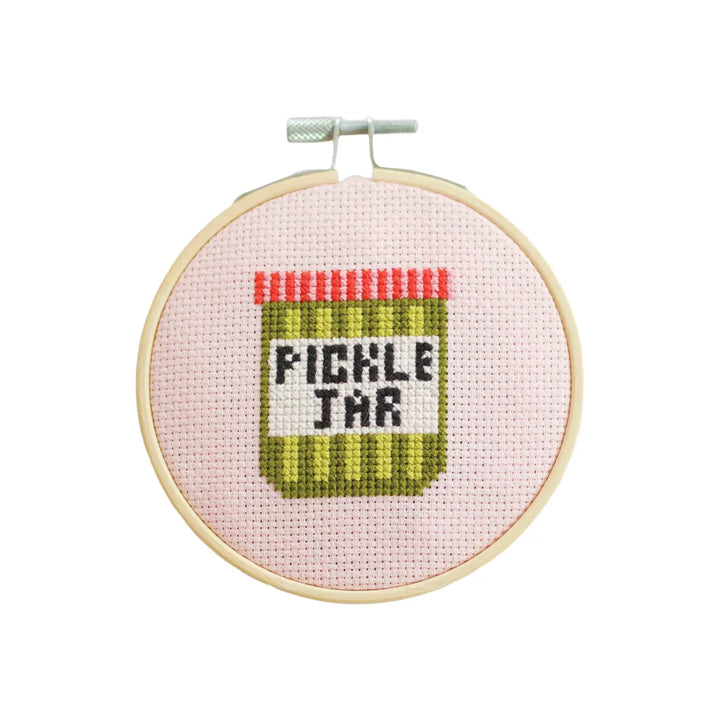 Cross-stitched pickle jar design on a pink background with a wooden hoop.