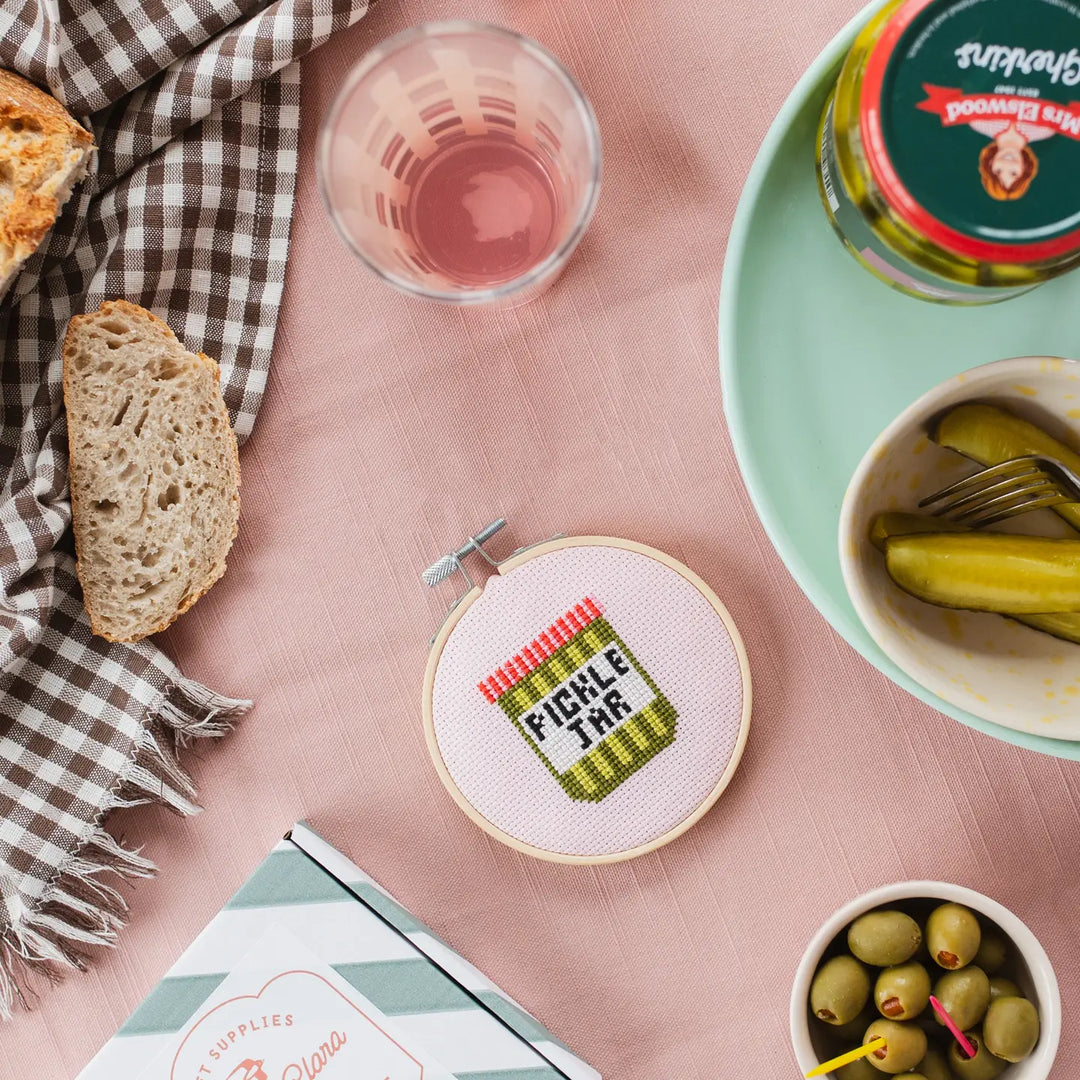 Cross-stitched pickle jar on a pink surface with bread, tea, and pickles.