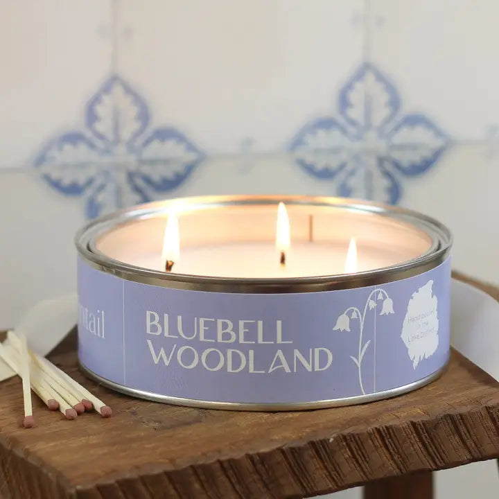 Candle in a 'Bluebell Woodland' tin with matches on a wooden surface.